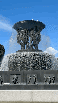 A large basin style fountain, mounted on a base. About the same height as two double decker buses, it's pumping water out continually, and the sheets of water are falling over the edges of the grey stone basin. The sky is blue, there are sculpted stone male figures standing on the base, holding up the basin. The splashing water creates white spray as it lands.