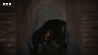 Claudia Winkleman, wearing a hooded cloak, slowly looks up to camera, brings a finger to her lips, and shushes.