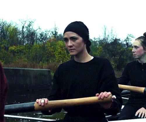 A young white woman with dark hair pulled back sitting in a competitive rowing boat holding an oar