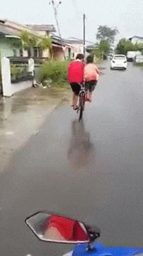 Video gif. Phone camera captures two children riding a bike down a wet residential streets, one standing on the left pedal and the other on the right pedal. Video gif. Phone camera captures two children riding a bike down a wet residential streets, one standing on the left pedal and the other on the right pedal.