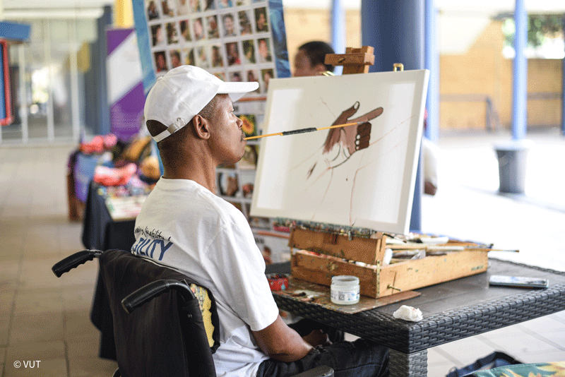 A Black disabled artist is painting a hand pointing to the right using a paintbrush in his mouth.