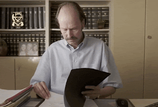 A plain looking man - middle aged, balding, wearing a beige shirt - is sitting at a desk reading a document. He looks up to the camera, slowly, and looks sceptical at what he's reading.