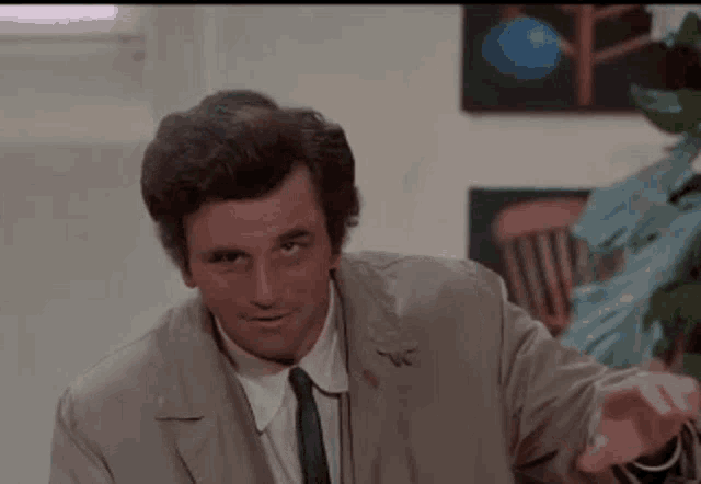 Gif dalla serie "Colombo". Il tenente Colombo, vestito con il suo classico impermeabile beige, sbatte una mano sull'altra e fa un cenno di assenso. Sullo sfondo si intravedono una finestra, una parete bianca e una pianta.