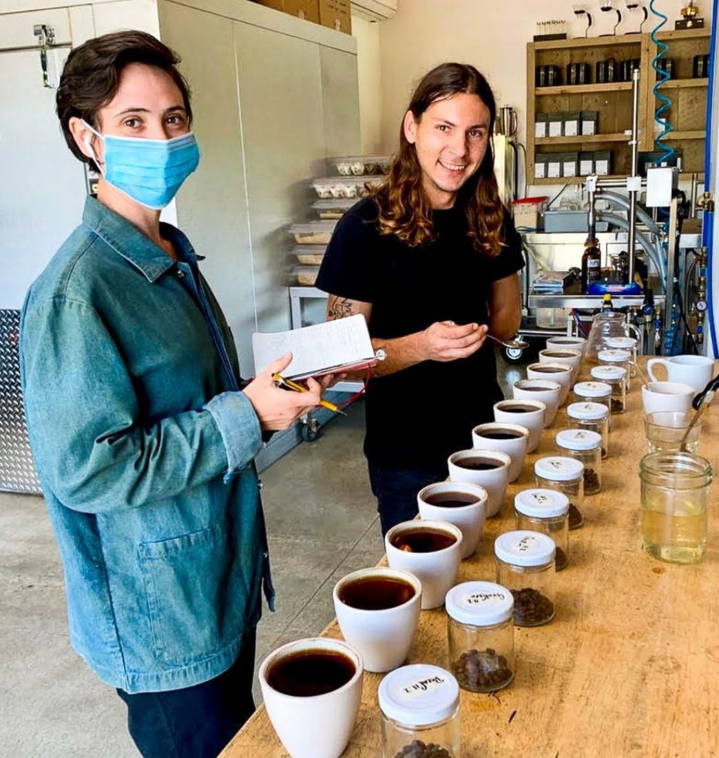 Siri (woman) and Joey (man) stand next to a coffee cupping table. A line of brewed coffee's stretch the length of the table waiting to be tasted. Siri has a notepad and pencil, and joey holds a tasting spoon.