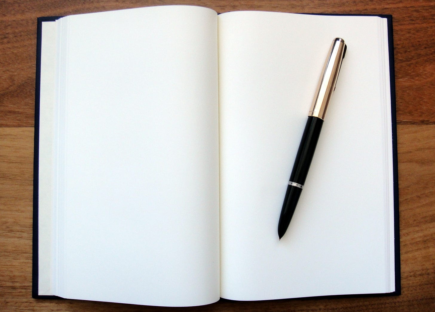 A black journal is open on a brown wooden table to blank white pages and a black and gold fountain pen lying on top. 