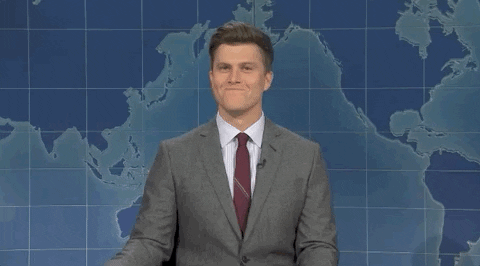 Colin Jost from SNL is at the news desk with a world map behind him. He is wearing a suit and slaps the desk as he tries to hold in a laugh. 
