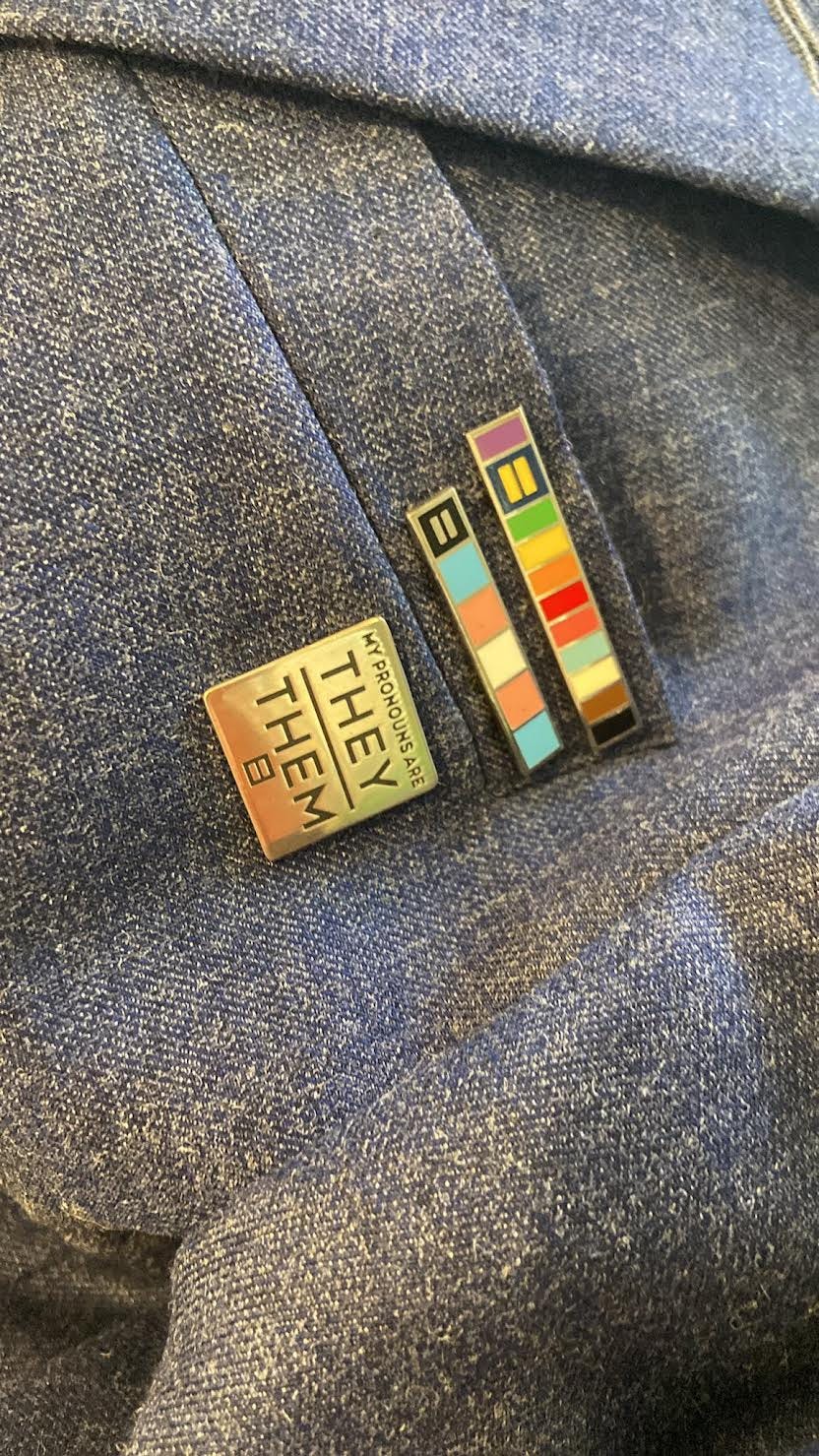 three pins; one with stripes of the progress flag colors, a second with stripes of the trans flag colors, and the third reading as "my pronouns are they/them"; all sporting the human rights campaign logo