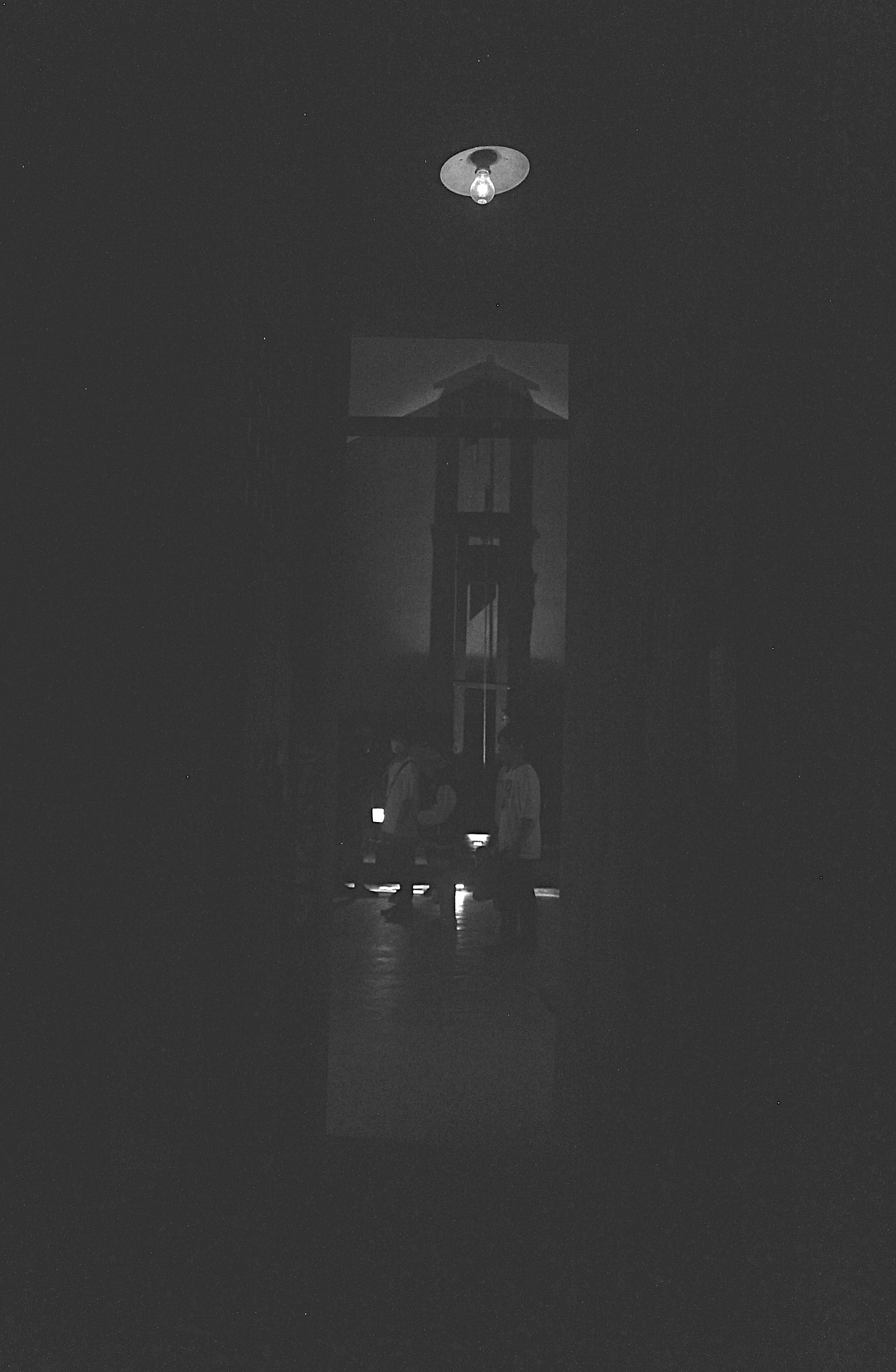 A black and white photo of a hallway that is lit by a single bulb. At the end of the hallways is the silhouette of a guillotine.