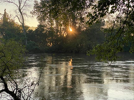 The first glint of light peeks out through the trees and reflects on the river. Photo by Dr. Jo.