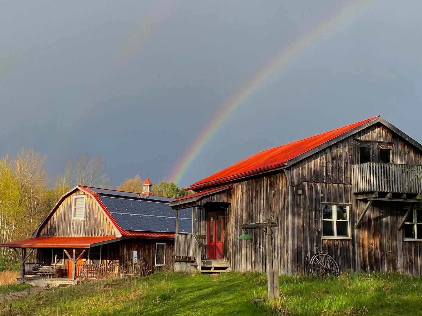 A double rainbow over two barn-like structuresr with red rooves. One roof has solar panels on it. 