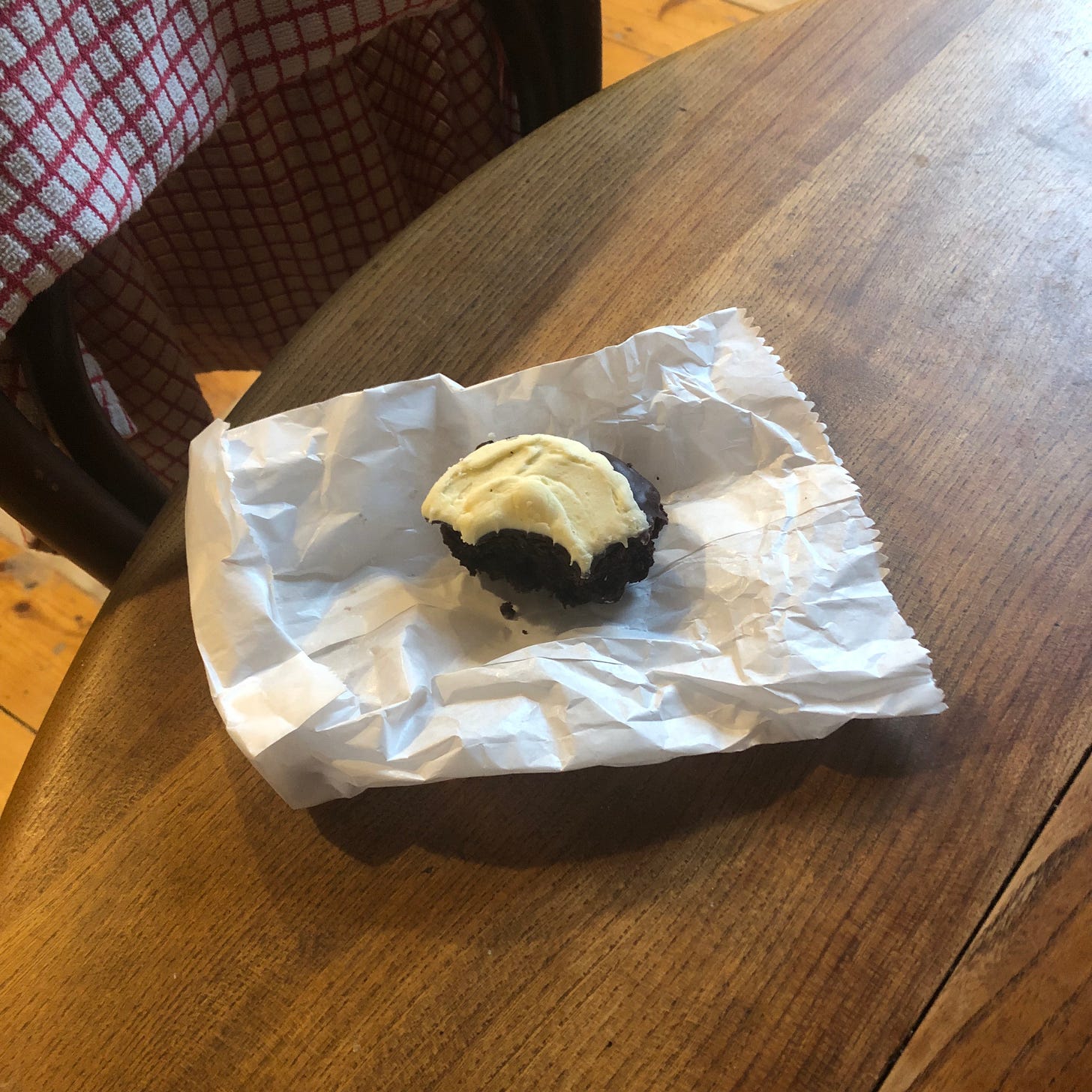  A photo of a half eaten chocolate brown cookie with white icing, laid atop a crinkled white paper bag. This sits on a brown wooden table, with a red and white gridded tea towel in the top left corner, resting against the back of a chair. 