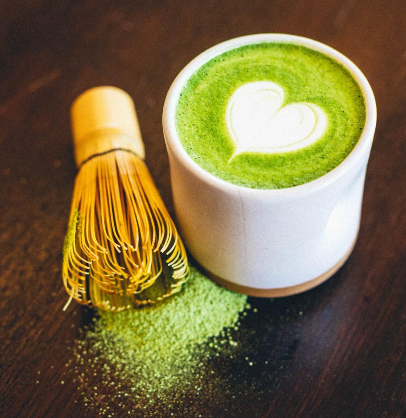A top view of a matcha green tea latte with a white heart latte art in the top. Drink is in a white ceramic cup with ground matcha tea sprinkled on a brown wooden table top.