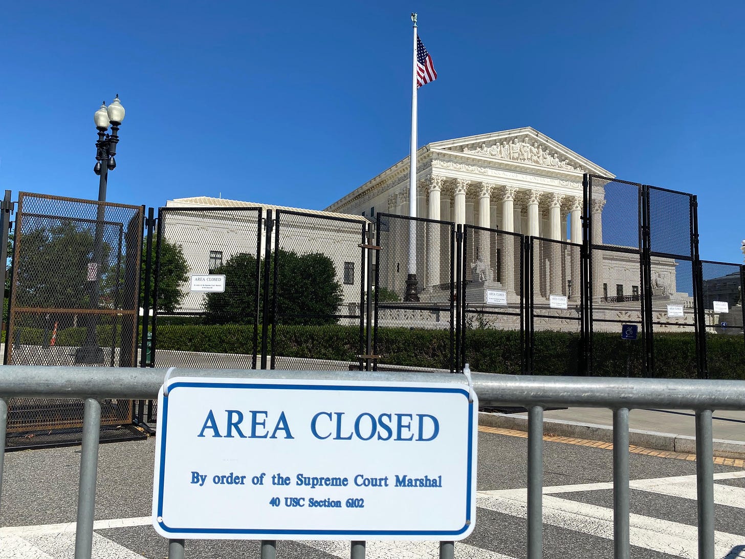 A photograph of the Supreme Court in the background. In the midground, a tall black fence that encircles the court grounds. In the foreground, another, lower, gray fence with a sign, "AREA CLOSED By order of the Supreme Court Marshal, 40 USC Section 6102.