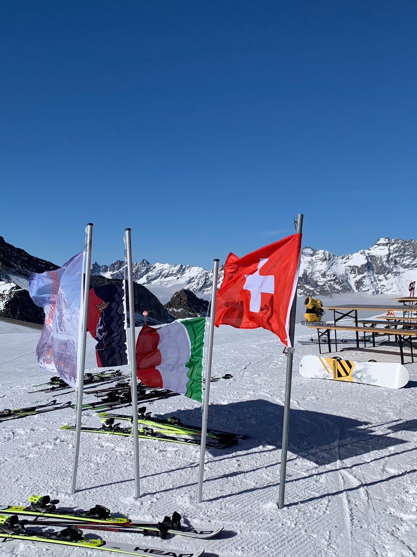 A photograph on a mountain peak with flags of Italian and Swiss provinces as well as the national flags of Italy and Switzerland. They are blowing in the wind with a mountain range behind them. Several pairs of skis and a snowboard lie on the snow and picnic benches from a restaurant refuge are visible on the right.