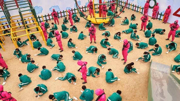 Scene from Squid Game where players are in a giant playground, bent over little cookies they're supposed to break a shape out of without damaging it. Guards loom ready to kill the losers.