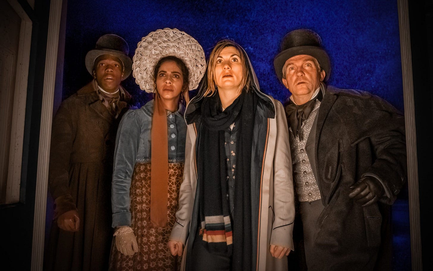 Team TARDIS--from left to right, Ryan Sinclair (played by Tosin Cole), Yasmin “Yaz” Khan (played by Mandip Gill), the Thirteenth Doctor (played by Jodie Whittaker), and Graham O’Brien (played by Bradley Walsh)--stand in the front entrance of the Villa Diodati. Ryan, Yaz, and Graham wear period attire, while all four people look offscreen at something that causes surprised expressions on their faces. Team TARDIS--from left to right, Ryan Sinclair (played by Tosin Cole), Yasmin “Yaz” Khan (played by Mandip Gill), the Thirteenth Doctor (played by Jodie Whittaker), and Graham O’Brien (played by Bradley Walsh)--stand in the front entrance of the Villa Diodati. Ryan, Yaz, and Graham wear period attire, while all four people look offscreen at something that causes surprised expressions on their faces.