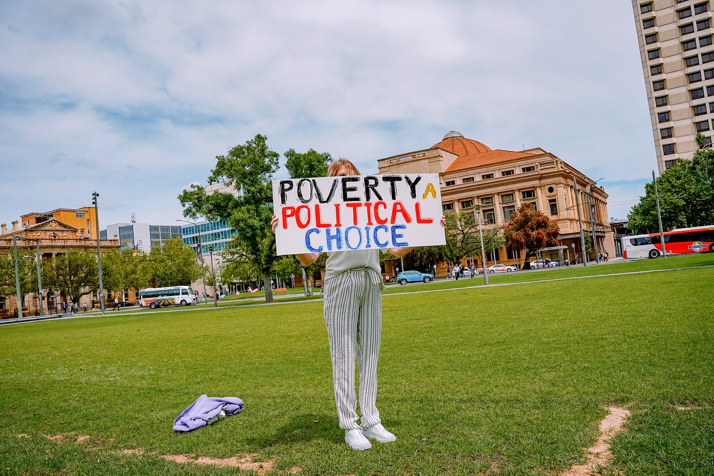 Woman holding a sign in a park with sandstone buildings in the background. The sign says "poverty is a political choice". 