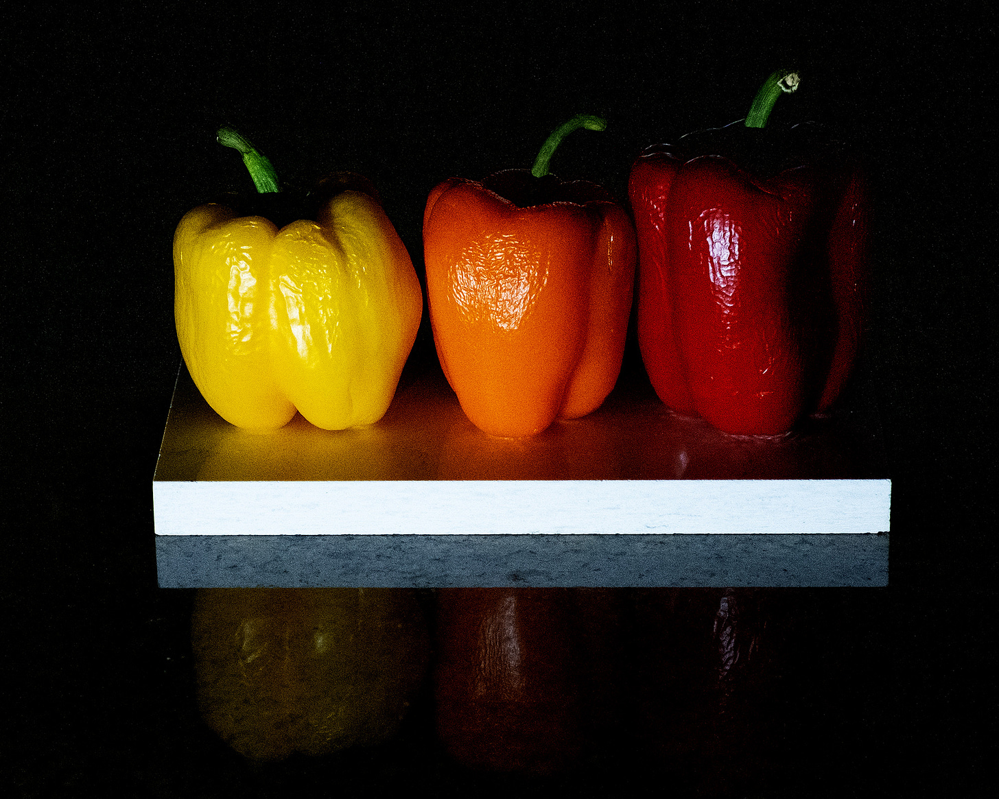 A red, orange, and yellow bell pepper arranged in a row on top of a white tile.  The scene is engulfed by shadows.