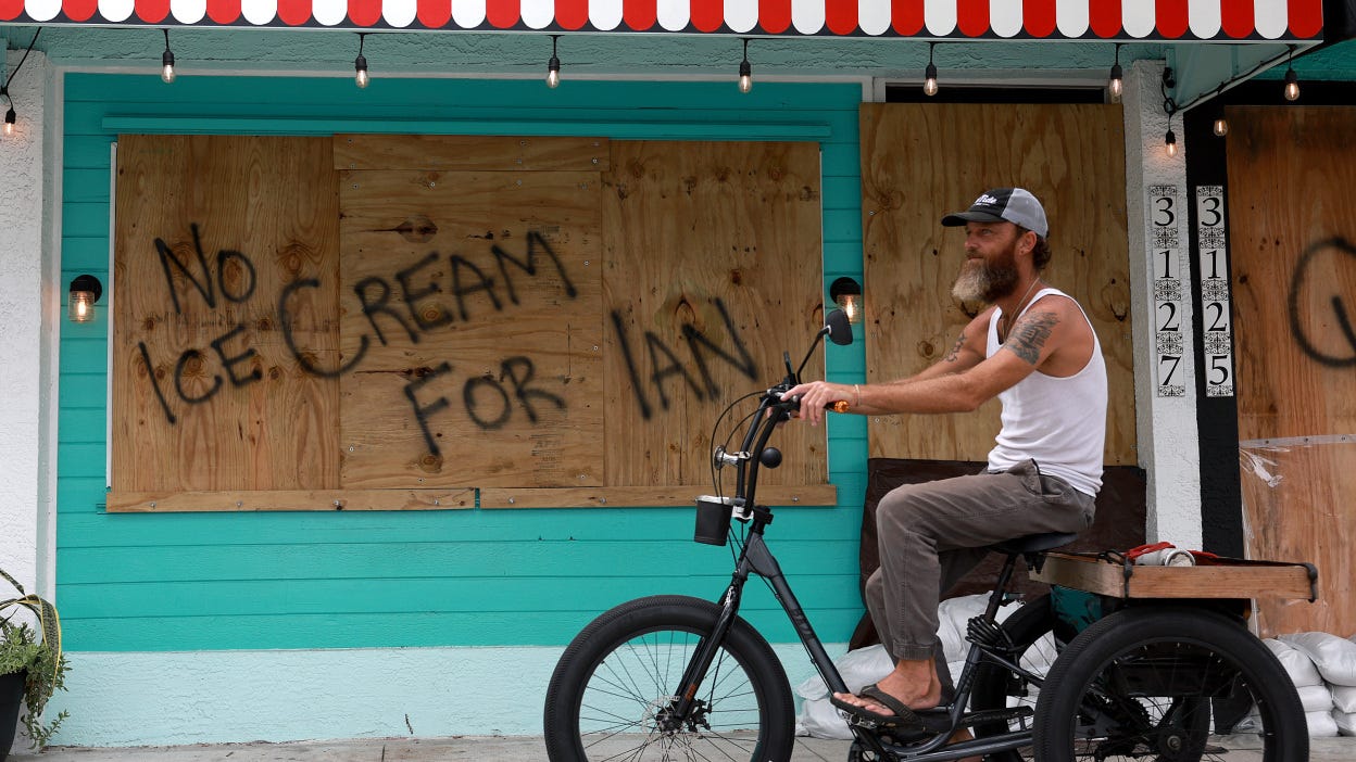 a man rides a bicycle in front of a boarded-up storefront upon which a shopkeeper has spray-painted "No ice cream for Ian."