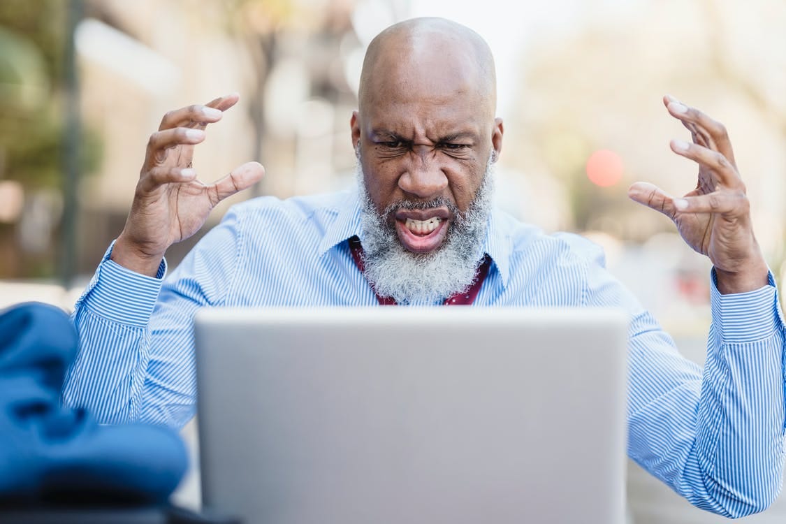 Wrathful focused bearded bald African American businessman in light blue shirt and tie watching laptop with raised hands while having video conversation