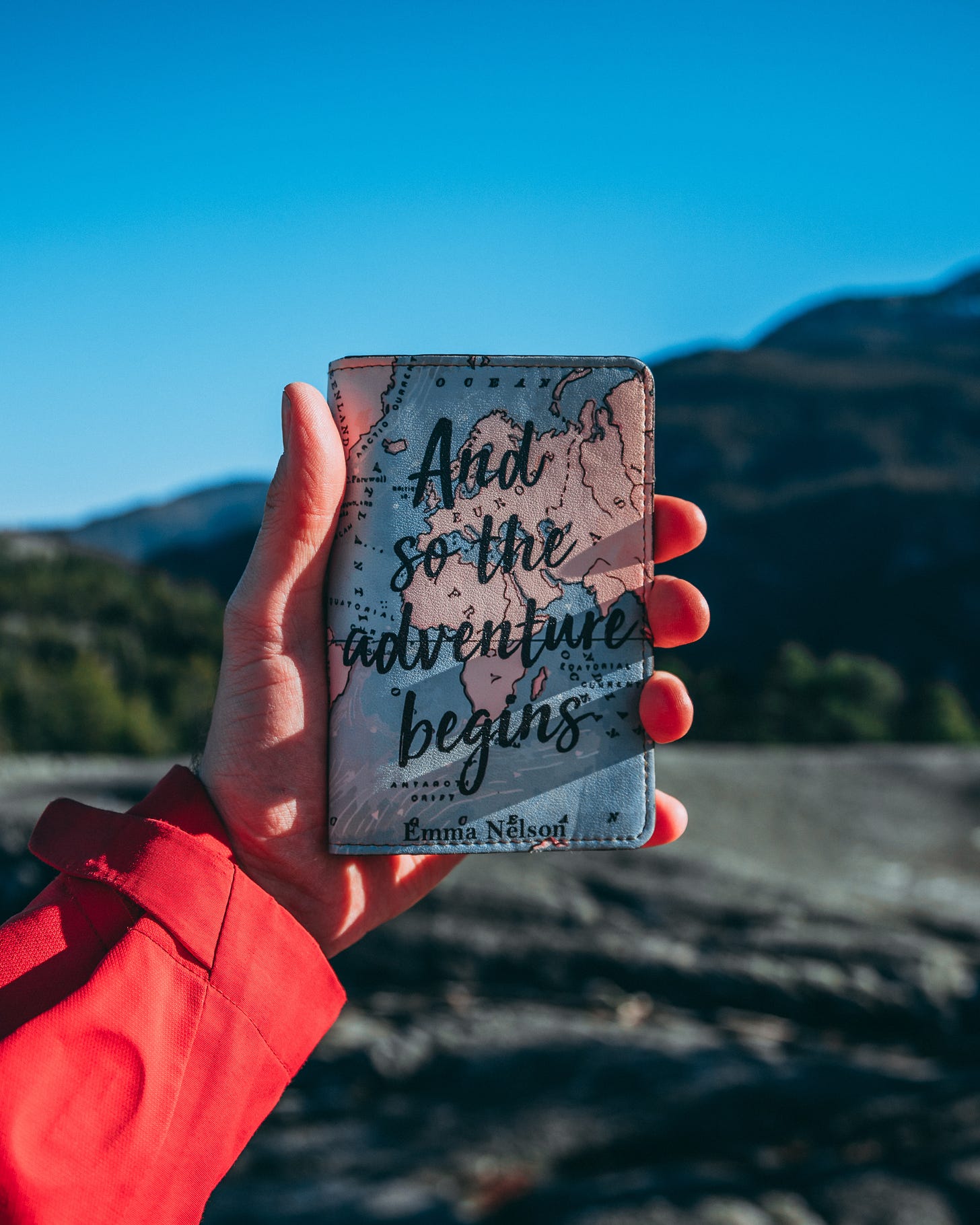 Mão segurando um passaporte com uma capa que diz "and so the adventure begins". Ao fundo há um cenário de montanhas e céu azul ao horizonte, a mão que segura é de uma pessoa branca e ela está com um casaco vermelho.