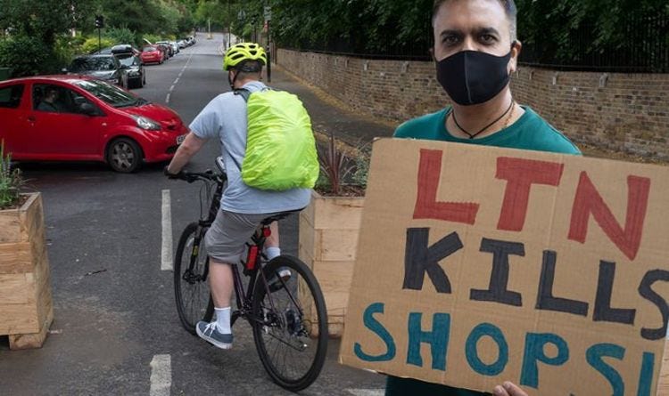 A cyclist passes by a planter. A car is turning in the background. In the foreground, a man with a face covering on holds a sign saying "LTN kills shops"