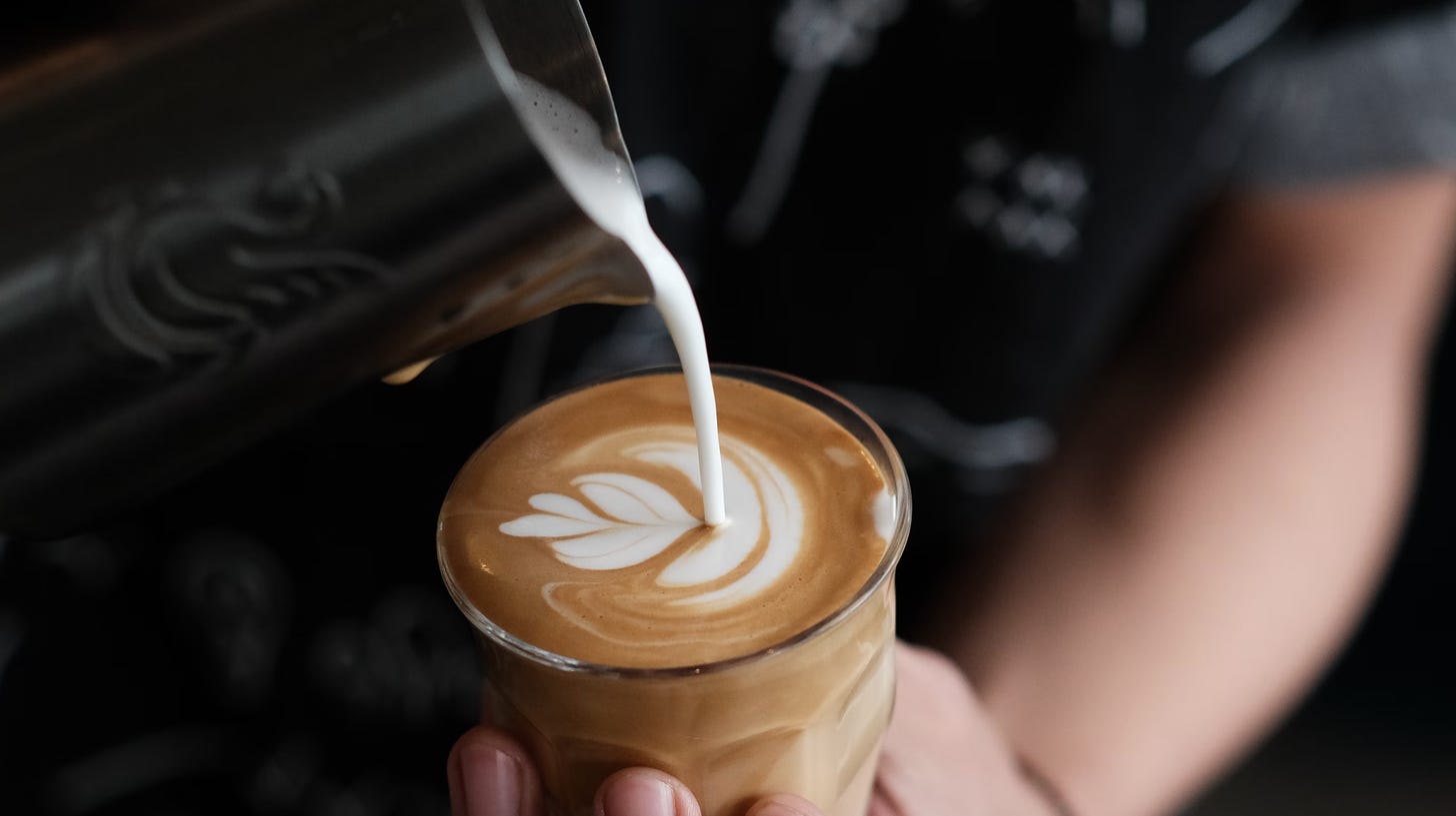 A close-up of steamed milk being poured from a silver pitcher into a glass coffee cup completing the heart shaped latte art on top of a cappuccino.