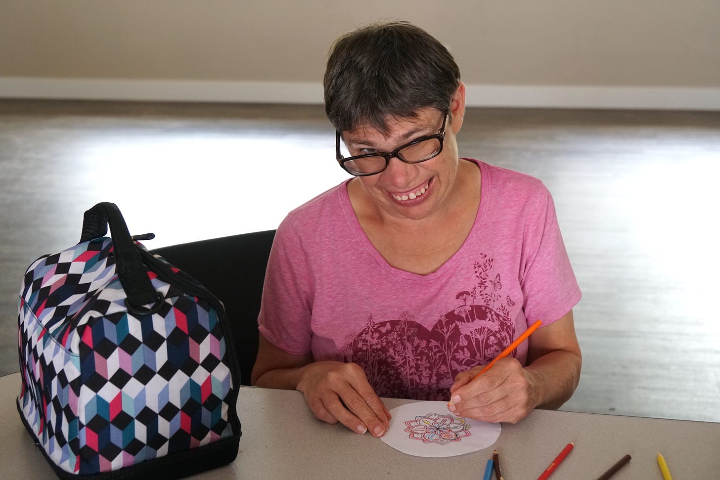 A woman with short dark hair wearing a pink t-shirt, smiles as she looks over the dark frames of her glasses. She holds an orange pencil in her left hand and is coloring a mandala which she holds on the table with her other hand.