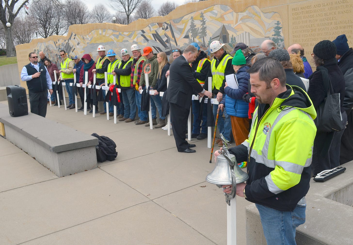 a line of people wearing neon vests and some hard hats stand behind white crosses in front of the workers monument mural