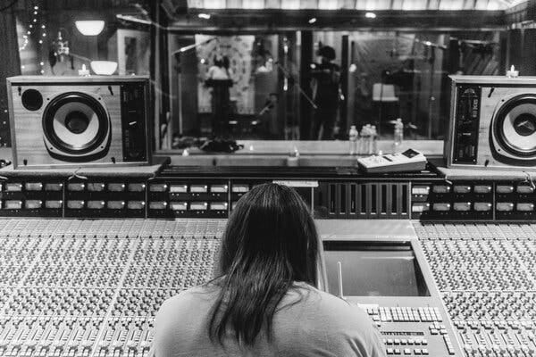 Martin sits at the recording console, with two singers visible in the distance, both standing in recording booths at microphones.