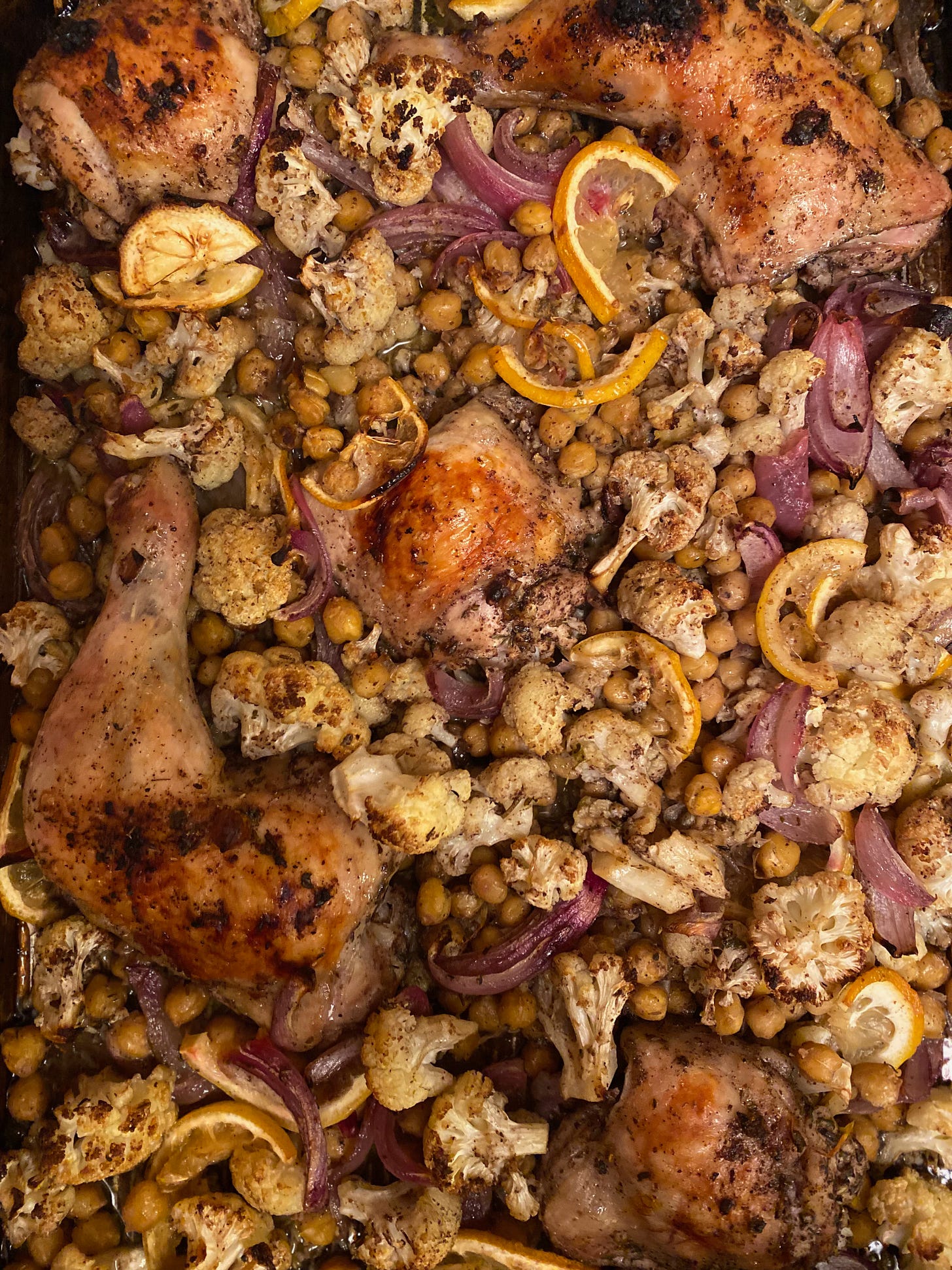 A close up of a tray of browned chicken things and breasts, lemon slices, charred cauliflower, red onion wedges, and chickpeas.