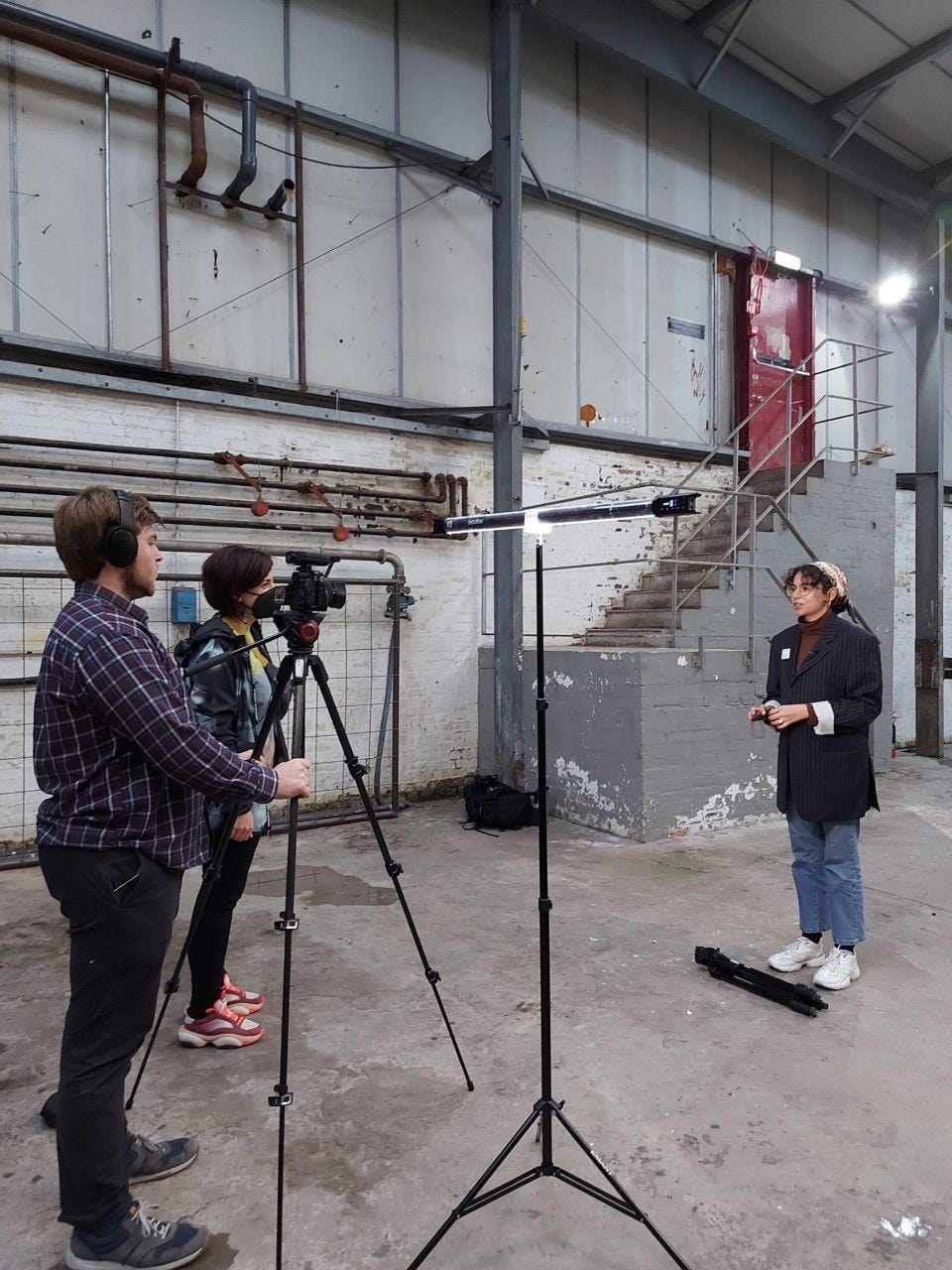 Hajar, a young Muslim woman in a black suit jacket and blue jeans, talks to a camera with two camera-people filming. They are in a warehouse.