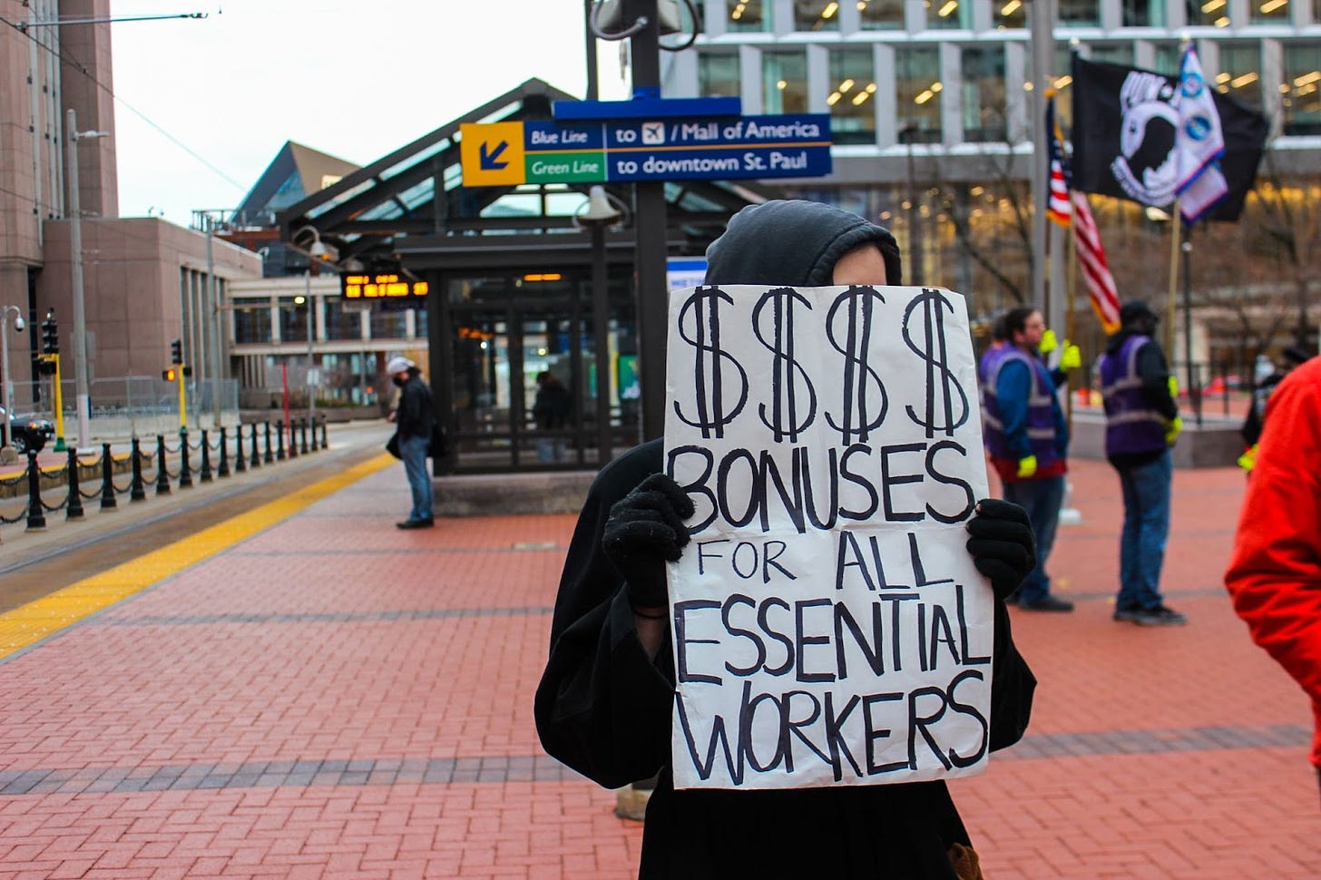 person in black hoodie standing in front of light rail station downtonw with a sign covering face reading "$$$$ bonuses for all essential workers"