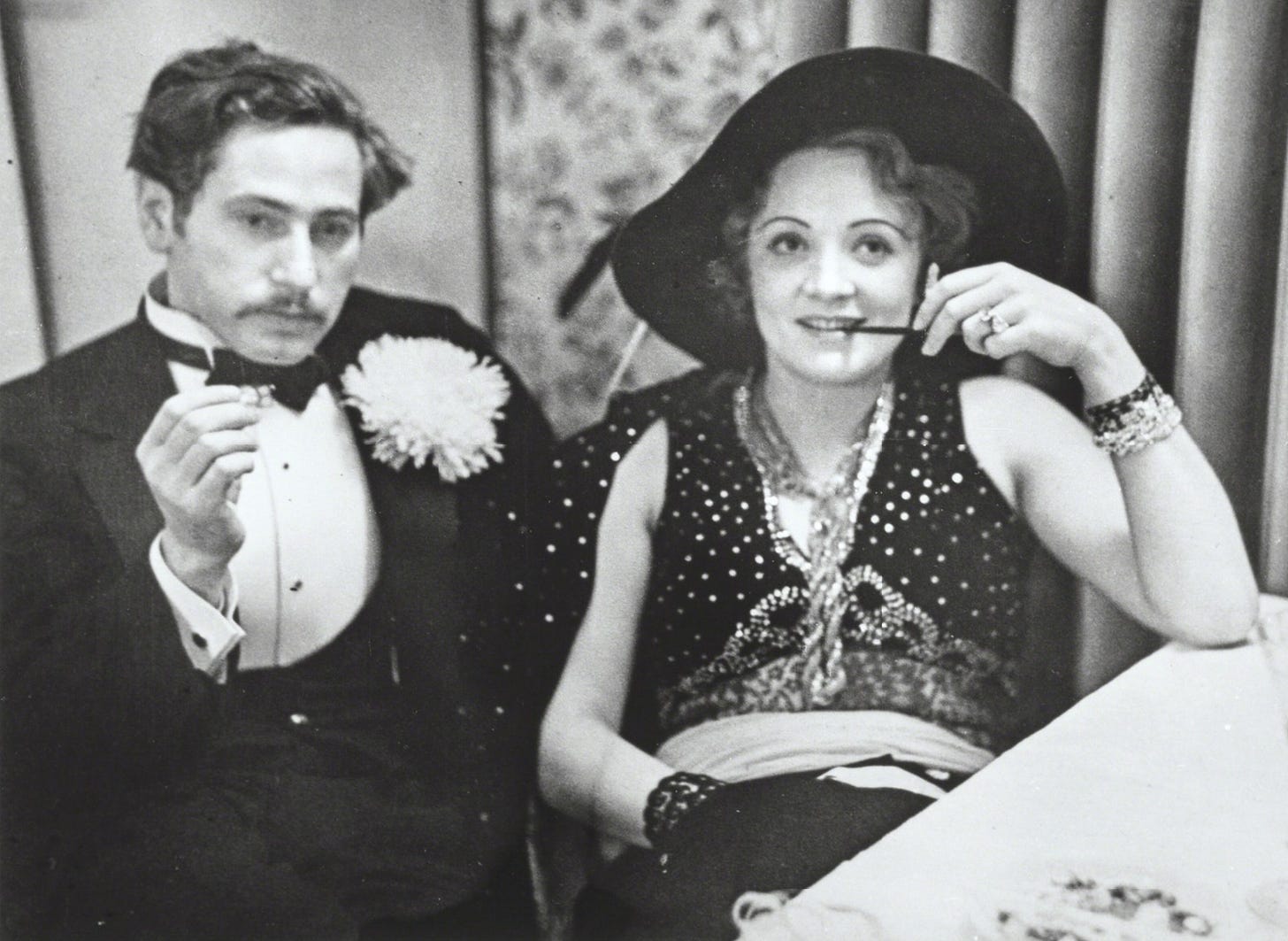 black and white photo of Josef von Sternberg in a tux, smoking a cigarette, sitting next to Marlene Dietrich with her pipe in hand