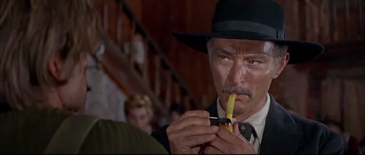 Mortimer (frame left), pipe in hand and blown-out match held up, frowns at Wild in frame left.