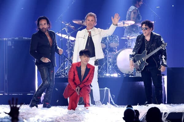 The members of journey arranged in front of its drum kit onstage: Marco Mendoza in black; Arnel Pineda, kneeling in a red suit; Jonathan Cain in a white jacket and pants; and Neal Schon in all black.