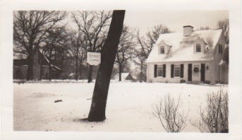 Black and white photo of a small white house and a front yard covered in snow. On a tree trunk in the foreground is a hanging sign that says "Riverby"