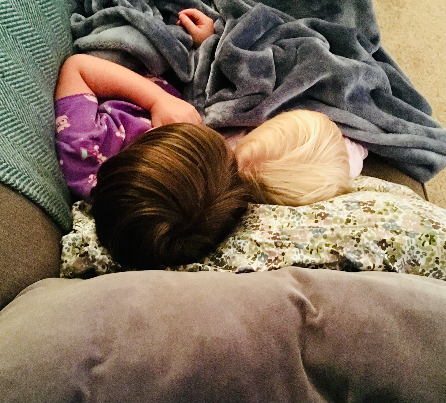 Two little girls laying on a couch with only the tops of their heads showing; one with brown hair, one with blond hair