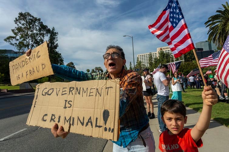 A man holds a sign saying 'government is criminal' and 'pandemic is a fraud' next to a child waving an American flag