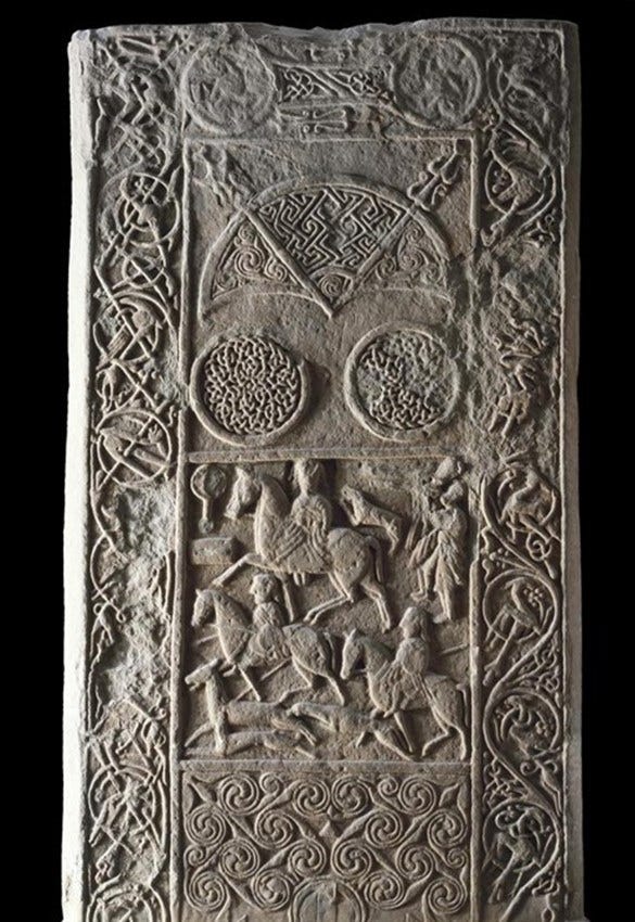 Photograph of the symbol-bearing face of the Hilton of Cadboll stone, with intricate relief carvings that include three Pictish symbols, a hunting scene and borders of ornate vine scroll.