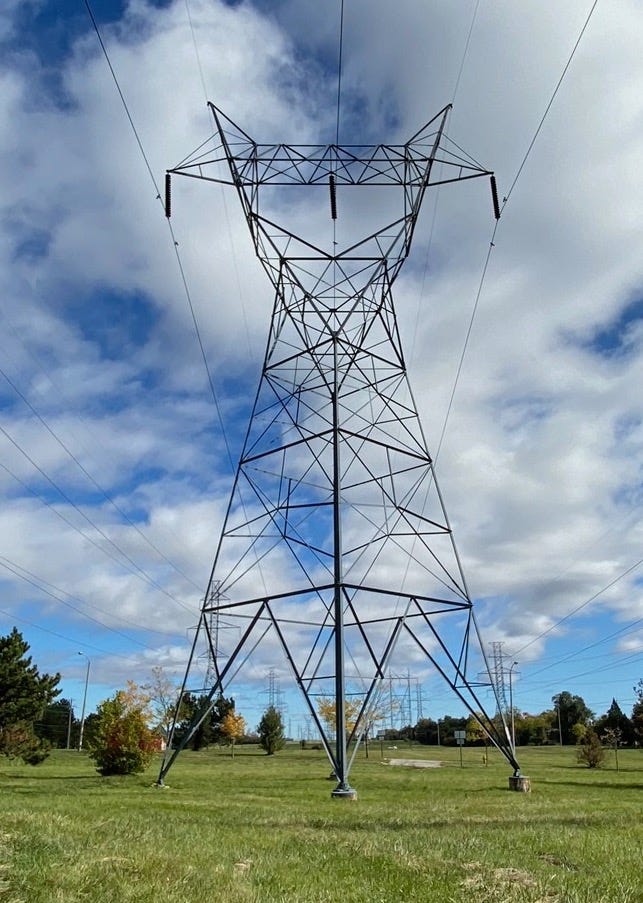A view of a metal transmission tower from the ground in Ontario, Canada, against a green field, rising into a clouded blue sky