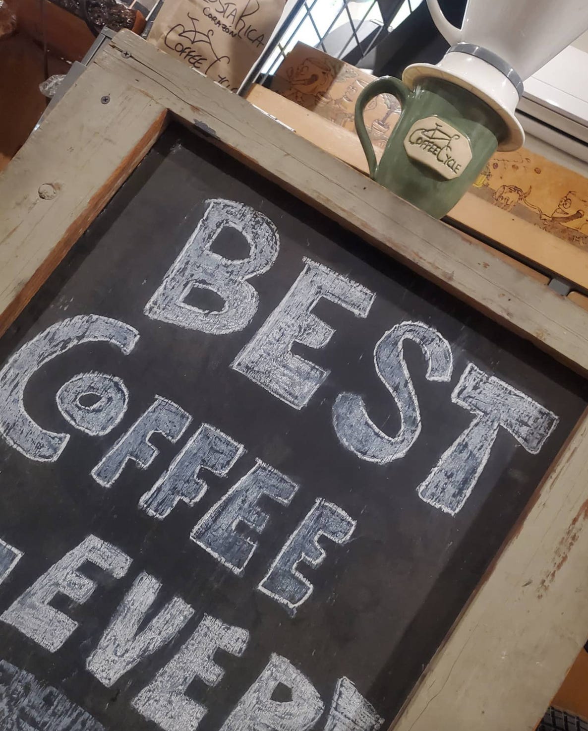 A close-up on the hand made wood a-frame chalkboard sign that sits in front of Coffee Cycle cafe. White chalk letters say "Best Coffee Ever!" A Coffee Cycle mug sits on top. The frame is made from upcycled pallet wood and painted light grey.