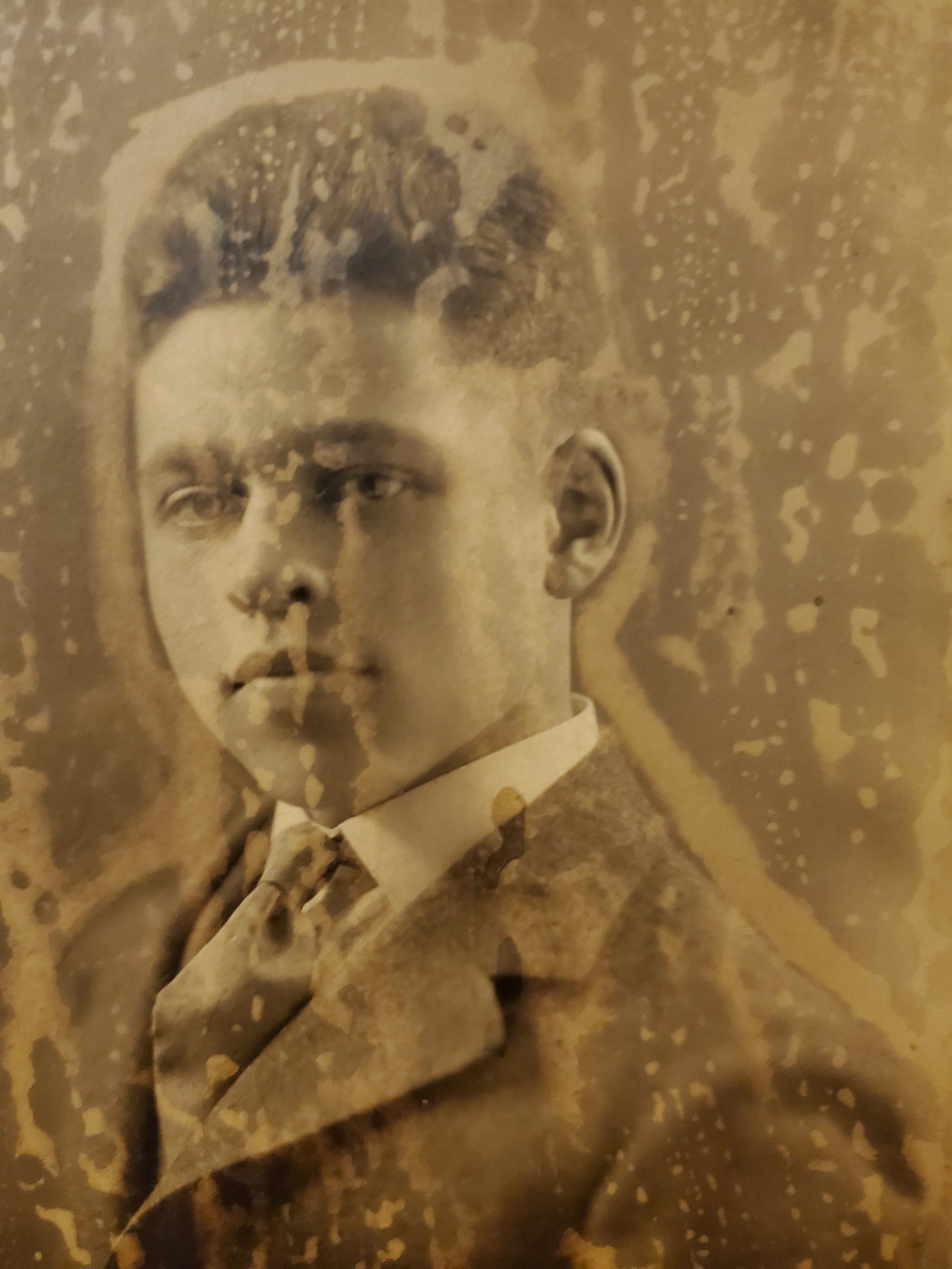 A damaged sepia photograph of a young man with short, curly hair, in a brown suit with a tie. 