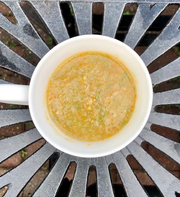 A white bowl is filled with yellow tomato gazpacho. It sits on a silver metal table.