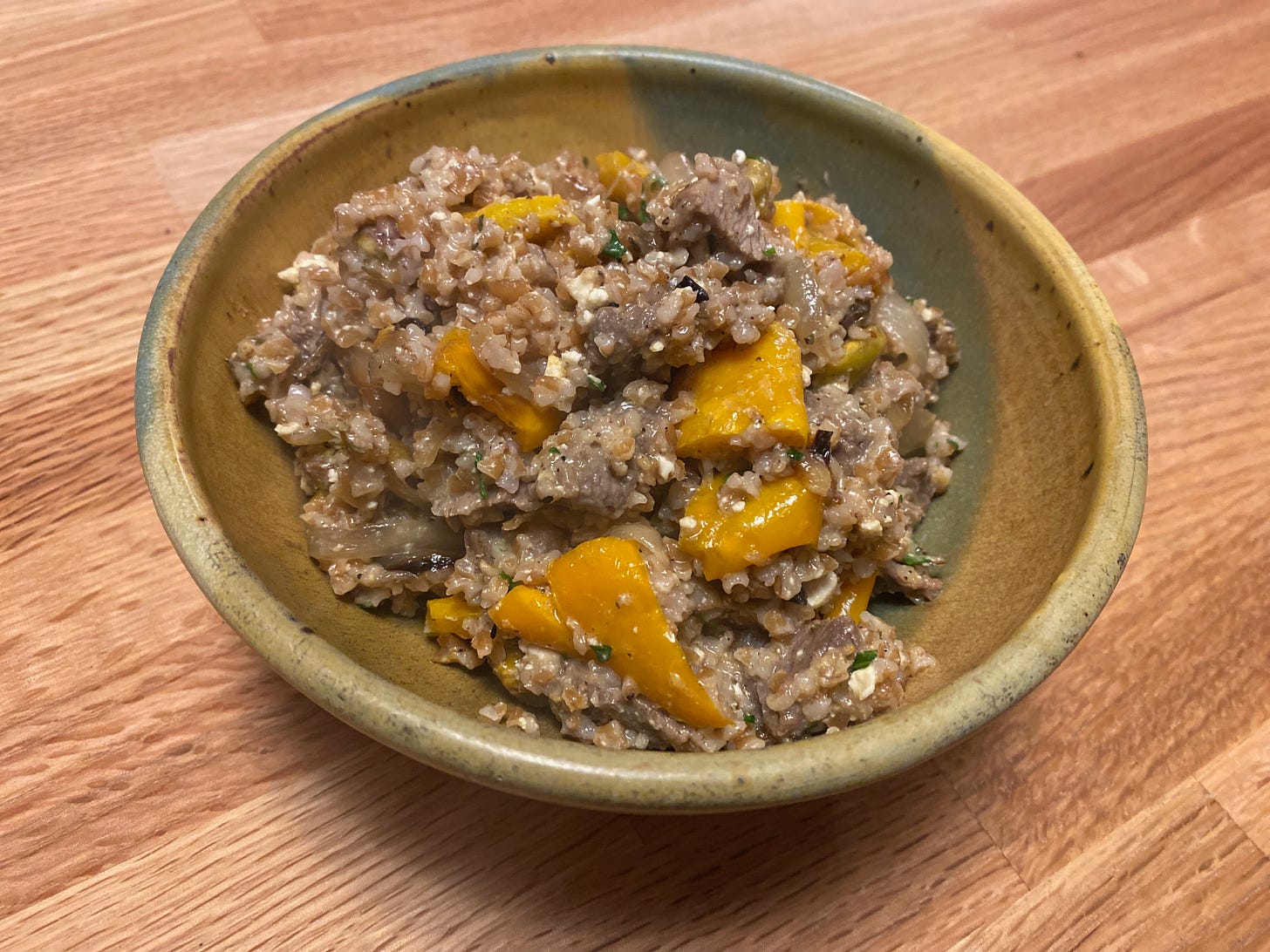 A ceramic bowl full of bulgar, bright yellow roasted peppers, and small chunks of browned lamb sits on a wooden counter.