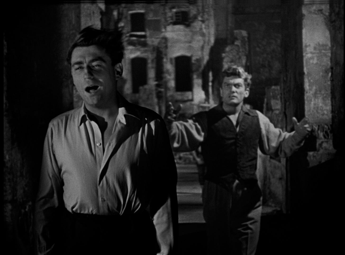 Black and white image. Two men walking with wind blowing in their faces. The front one is making a kind of wry comment with mouth wide open and wearing a shirt tucked into high-waisted trousers, 50's style outfits. The film was made in 1950. The man behind is tall, handsome, frightened looking and throwing both hands up by his sides, as if confused and grasping for meaning. They are passing through an old Grecian looking corridor, with ramshackle houses with black, paneless windows and ivy creeping up and down.