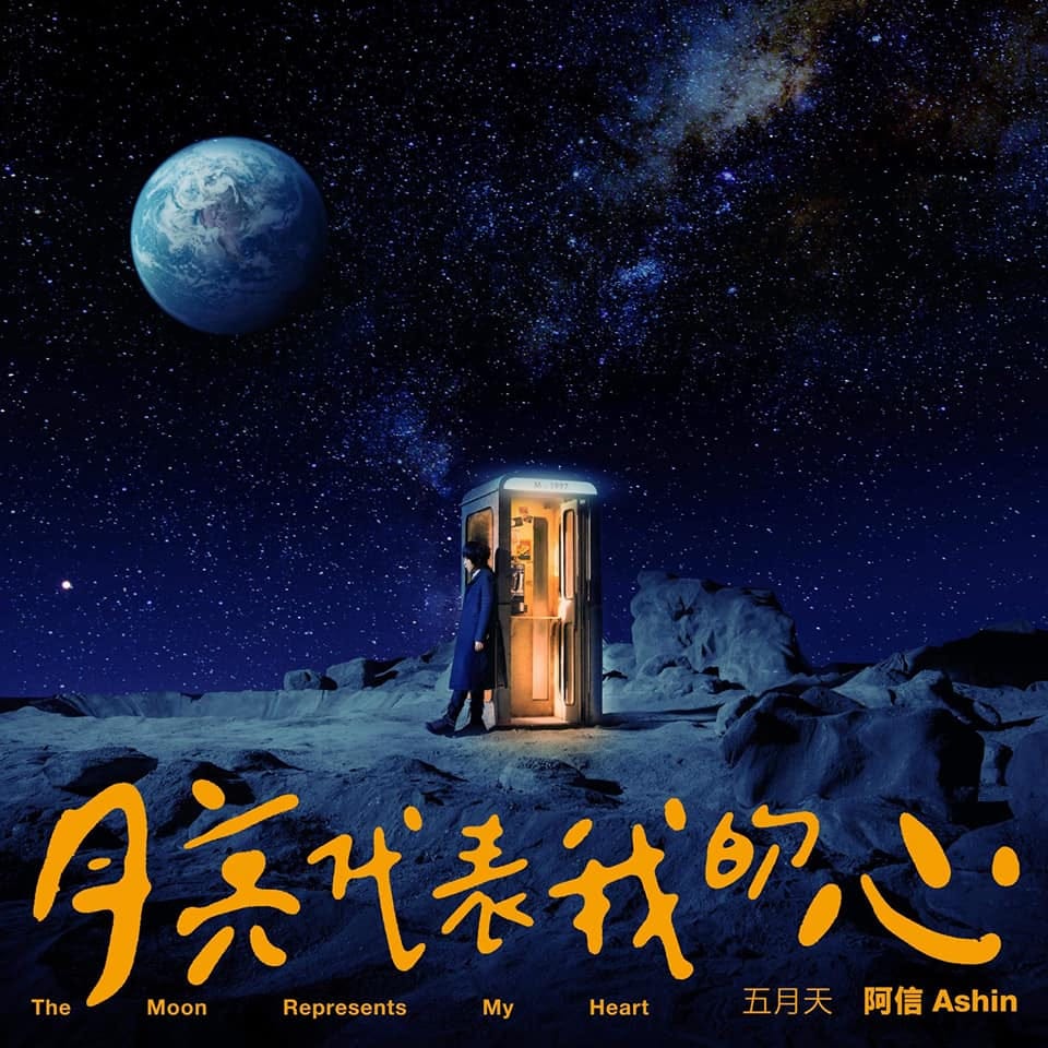 A man wearing a long blue coat stands on a moonscape in front of a warmly lit phone booth in the middle ground. In the background the background, the Milky Way and the Earth are visible. In the foreground is the title of the song "The Moon Represents My Heart" in gold lettering. A man wearing a long blue coat stands on a moonscape in front of a warmly lit phone booth in the middle ground. In the background the background, the Milky Way and the Earth are visible. In the foreground is the title of the song "The Moon Represents My Heart" in gold lettering.