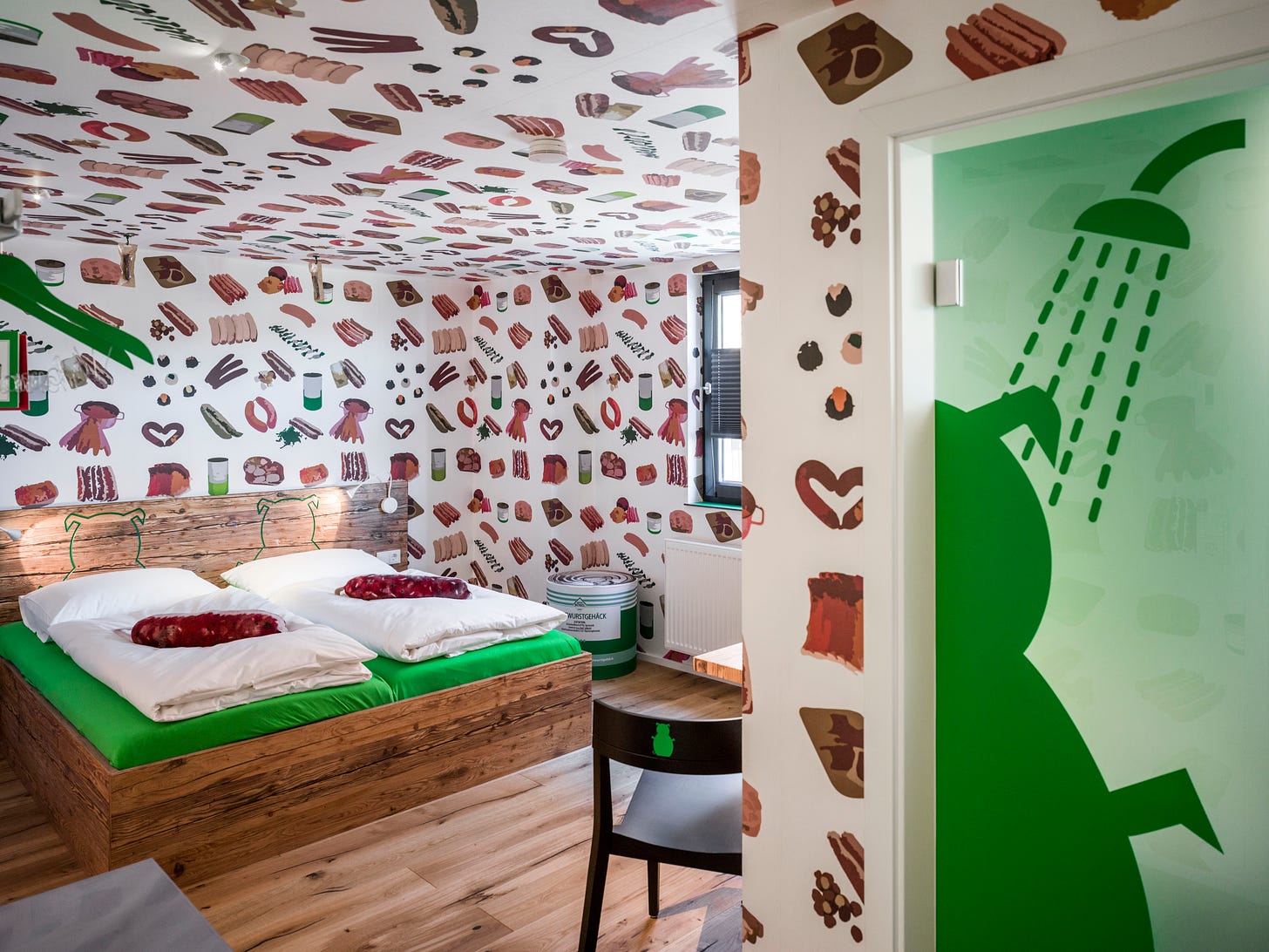 A hotel room with pictures of various sausages all over the walls and ceiling. There is a double wooden bed with bright green sheets and the bathroom door has a picture of a green pig in a shower on it.