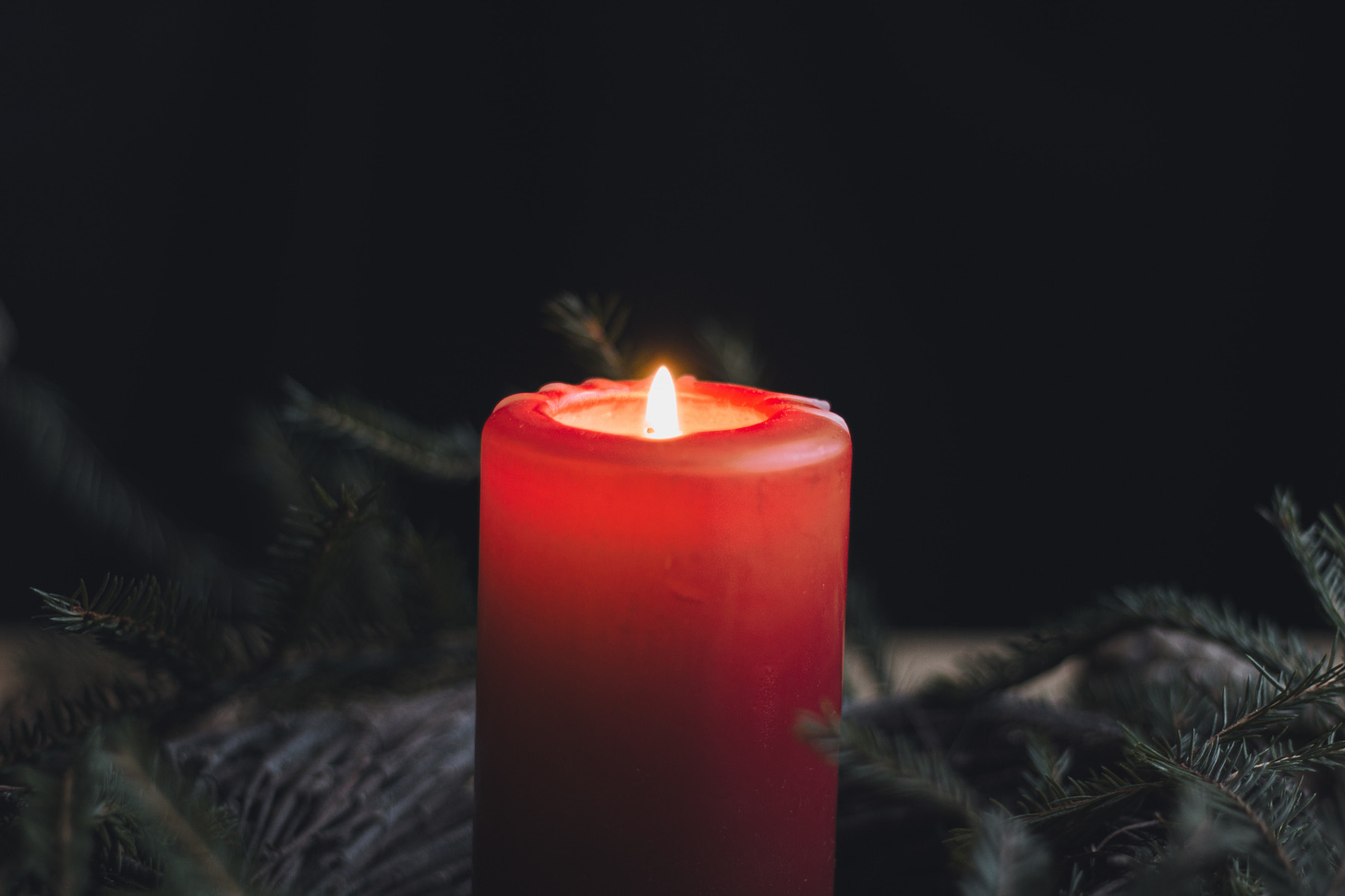 An illuminated red candle is at the centre of the image surrounded by fir tree branches against a black background.