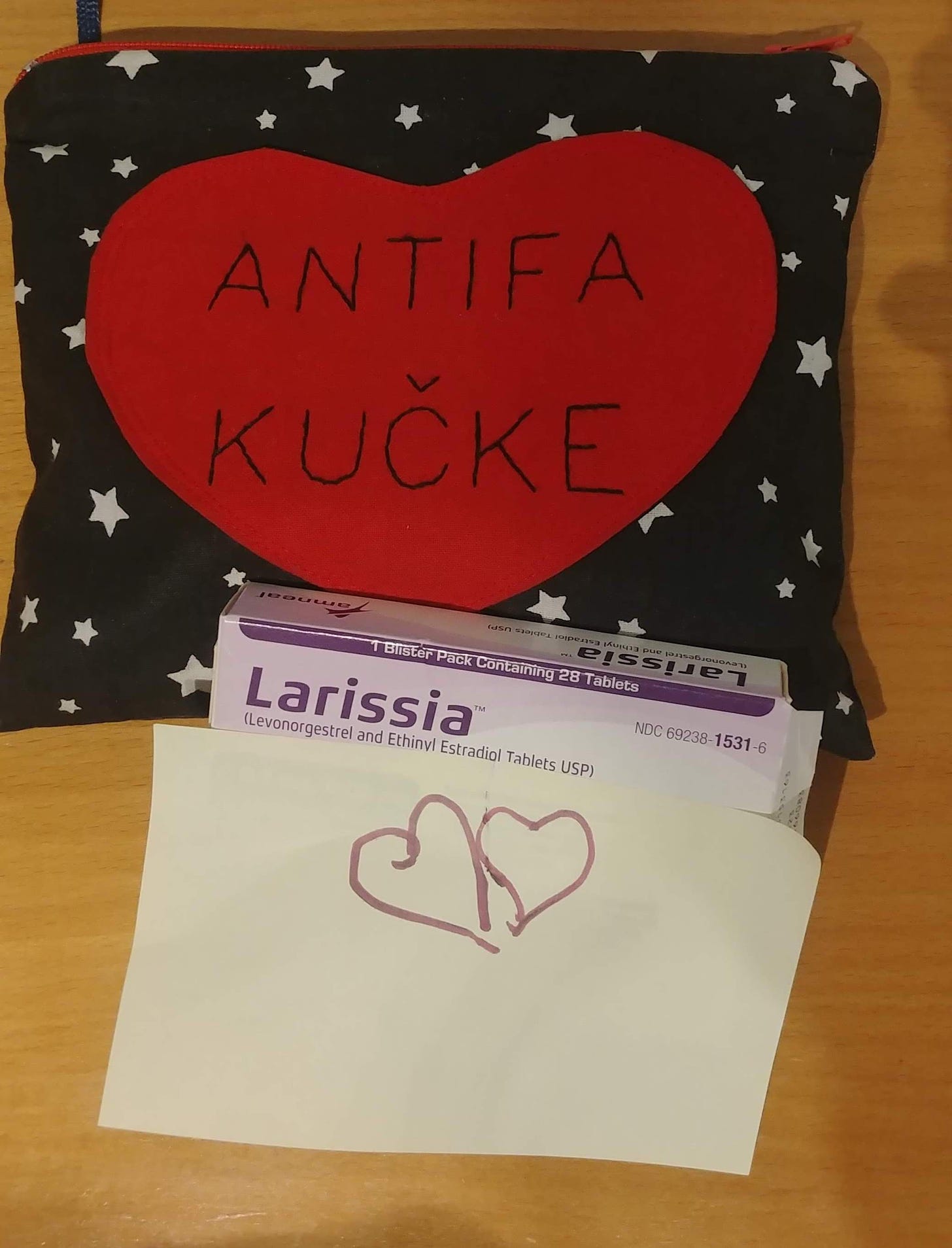 A photo of a table with several objects. At the top is a pencil case made out of black fabric with white stars on it. The front of the pencil case has a heart with "Antifa Kucke" stiched onto it. Below the pencil case is a box with Larissa, a type of birth control. Identifying details on the box are covered with a post-it that has a heart drawn on it.